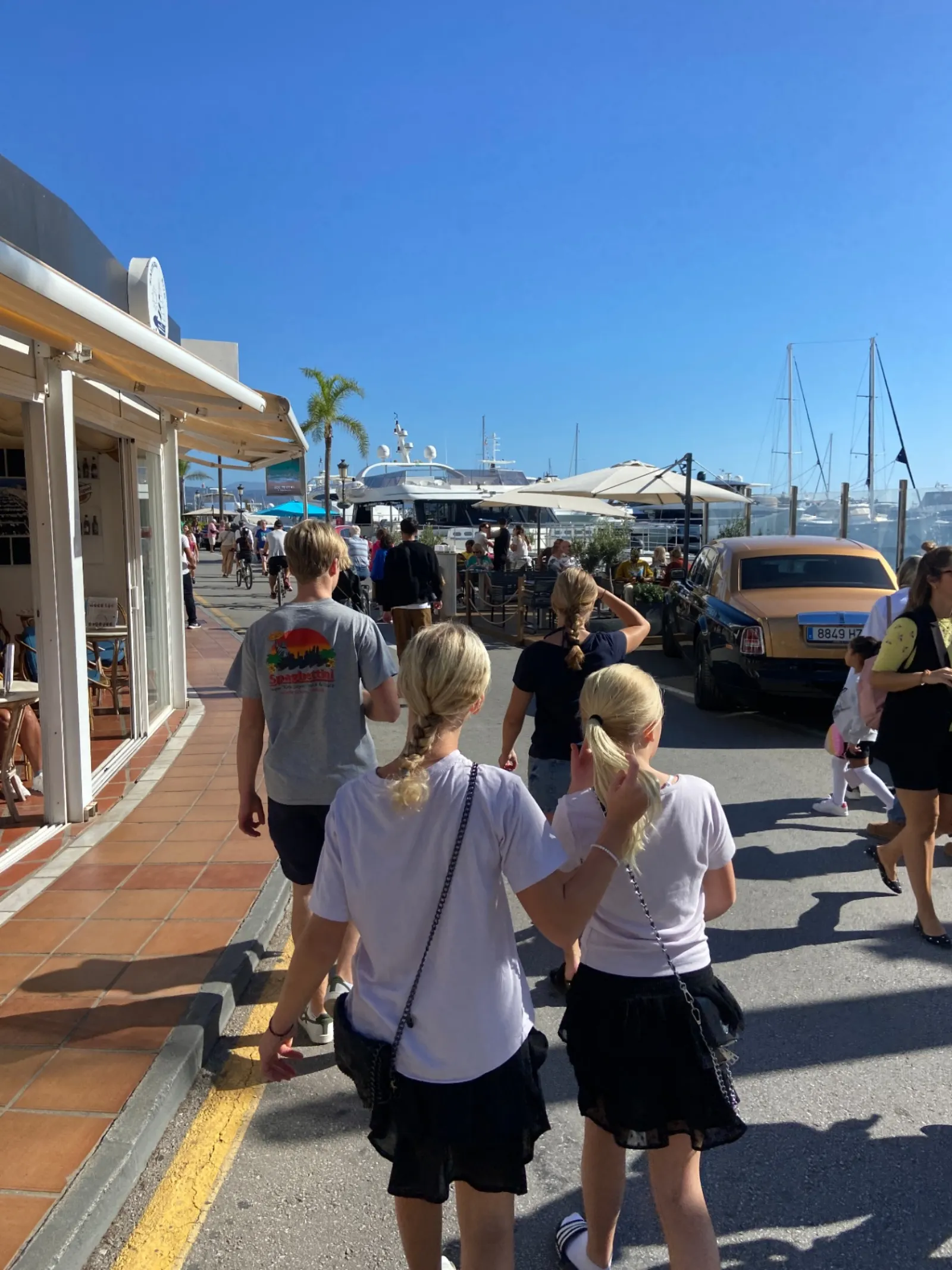 Visitors walking along the Puerto Banús marina promenade past yachts and a Rolls-Royce